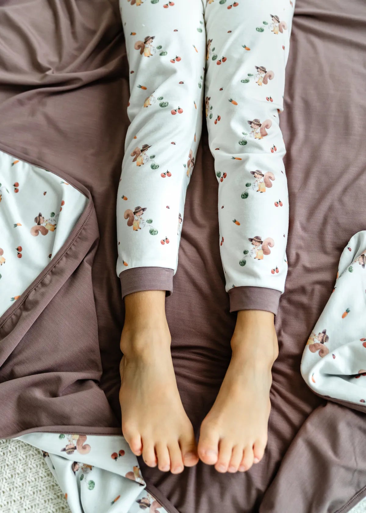 Child wearing pyjama pants with a squirrel watering vegetables pattern, lying on a bed with matching brown blanket