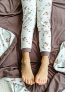 Child wearing pyjama pants with a squirrel watering vegetables pattern, lying on a bed with matching brown blanket