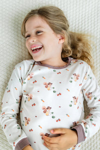 Young girl lying on a light coloured bed wearing white pyjamas with brown trimmings and a farmer squirrel pattern watering vegetables.