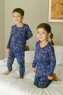 Two children in matching blue pajamas standing on a bed in a bedroom.