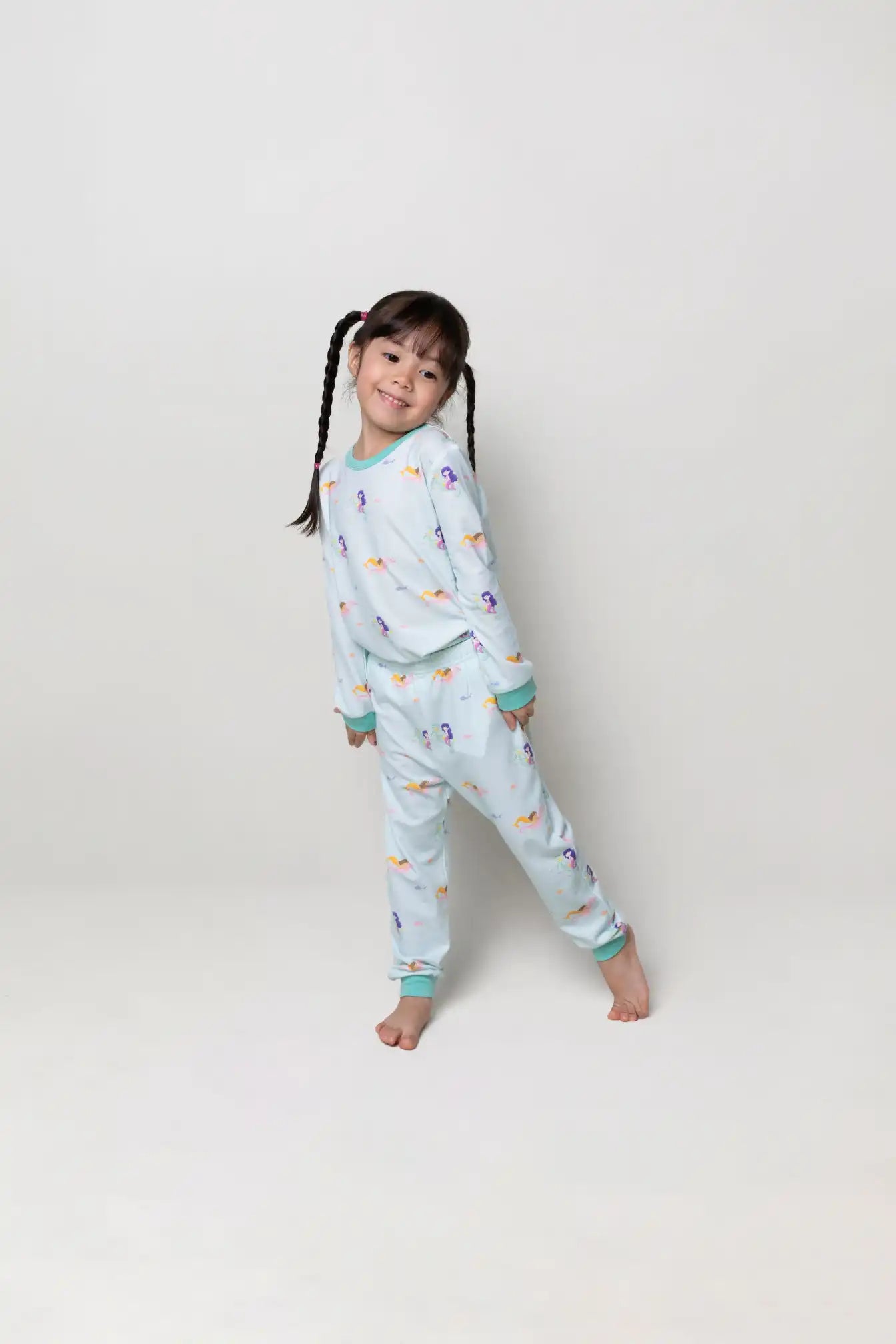 Child wearing a light turquoise pajama set with colourful mermaid prints on a white background and smiling.